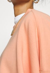 Sweatshirt couleur pêche avec une coupe décontractée, texture en molleton doux, col côtelé et coutures d'épaules tombantes. Présente des bijoux en chaîne dorée.