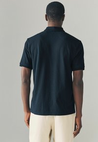 Polo negru cu un model geometric texturat, mâneci scurte și guler. Purtați cu pantaloni de culoare deschisă, evidențiind o croială lejeră.