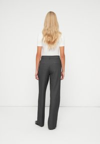 Femme aux longs cheveux blonds portant un haut blanc à manches courtes et un pantalon large gris sur mesure, debout de dos sur un fond blanc.