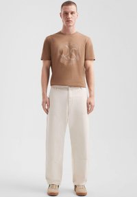 Brązowy bawełniany t-shirt z okrągłym graficznym logo, połączony z luźnymi, jasnobeżowymi spodniami i brązowymi sneakersami. Płaska faktura, minimalne akcenty.