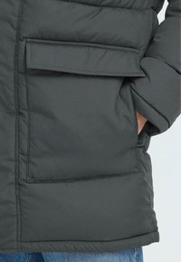 Mørkegrå pufferjakke med tekstureret overflade, der har en stor klaffelomme og et isoleret design for varme.