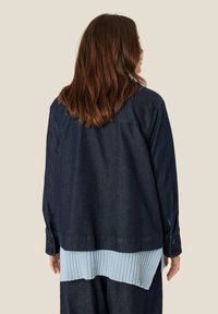 Mørkeblå denimjakke med en ribbet lys blå strikket underlayer, der har knaplukning ved ærmerne og en afslappet pasform. Vist fra bagsiden.