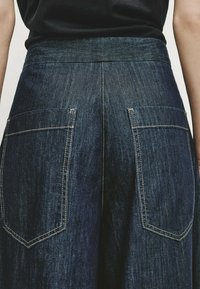 Vue arrière d'une personne portant un pantalon en denim bleu foncé avec de grandes poches arrière rectangulaires et des coutures visibles.