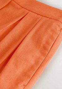 Gros plan sur un pantalon en tissu orange montrant la ceinture, les plis devant et les détails de la couture latérale sur fond blanc.