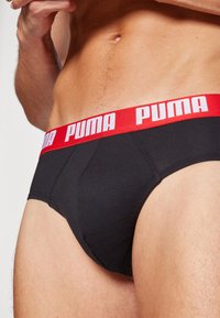 Sorte bomuldsunderbukser med en rød elastisk talje, der har "PUMA" i hvid skrift. Designet inkluderer en kontureret lomme og sømløse sider.