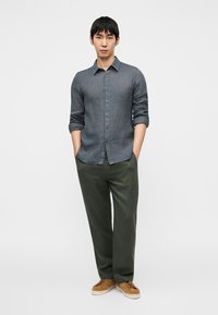 Γκρι λινό πουκάμισο με κούμπωμα με κουμπιά και μακριά μανίκια, συνδυασμένο με φαρδιά ελιά πράσινα παντελόνια και καφέ αθλητικά παπούτσια.