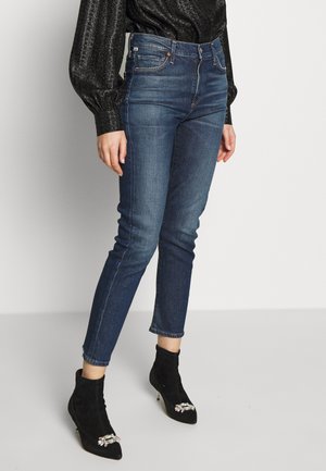 Kobieta w niebieskich, obcisłych, wysokowzrostych dżinsach, czarnej, błyszczącej bluzce z długim rękawem oraz czarnych botkach na obcasie z ozdobami w formie klejnotów.