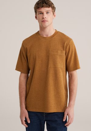 Bruin, T-shirt met korte mouwen en een gestructureerde afwerking, voorzien van een borstzakje. Ronde halslijn en een losse pasvorm. Gecombineerd met donkerblauwe spijkerbroek.