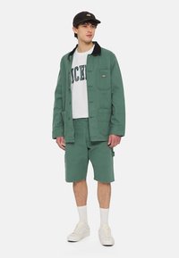 Zelená bavlněná bunda s černým límcem, bílý tričko s textem "DICKIES", odpovídající šortky a bílé tenisky.