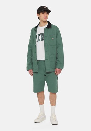 Chaqueta de algodón verde con un cuello negro, camiseta blanca con el texto "DICKIES", pantalones cortos a juego y zapatillas blancas.