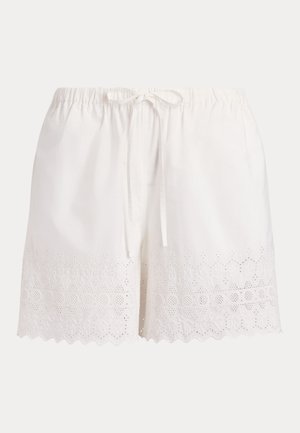 Žena v bielej blúzke z bielej dierkovanej čipky a šortkách, hnedý tkaný klobúk, hnedé sandále, drží zeleno-bielu pruhovanú tašku.