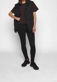 Černá prošívaná bezrukávová vesta přes tmavě hnědé tričko, v kombinaci s černými legínami a černými teniskami. Hladká textura a přiléhavý design.