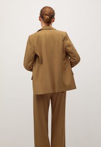 Blazer marron sur mesure à coupe décontractée, grands revers et pantalon à jambes larges assorti. Texture lisse, design minimaliste, sans accents visibles.