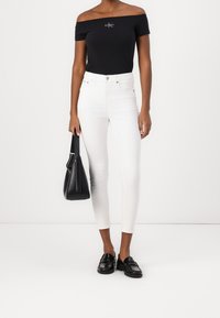Top negro sin mangas con logo, jeans skinny blancos, mocasines negros y un bolso negro. Textura suave y diseño ajustado.