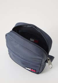 Tmavě modrá látková taška se zipovým otvíráním, jednou přední kapsou a malou nášivkou s logem. Šestiúhelníkový design s hladkým povrchem.