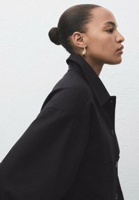 Giacca nera oversize con colletto e chiusura a bottoni, realizzata in tessuto liscio e con una forma strutturata. Il modello indossa orecchini a goccia d'oro.