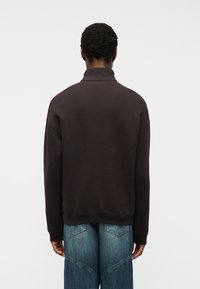 Άνδρας με πλάτη, φοράει σκούρο καφέ μπλουζάκι με μακριά μανίκια και μπλε τζιν φαρδιά παντελόνια, μπροστά σε σκηνικό απλής φωτεινής επιφάνειας.