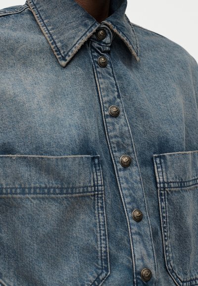 Τζιν μπουφάν με μεταλλικά κουμπιά με πατητήρι και δύο τσέπες στο στήθος, με κοντινή εικόνα του γιακά και του άνω μπροστινού μέρους.