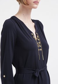 Marinblå klänning med lång ärm i slät tyg, med V-ringning och detalj av guldkedja samt knytning i midjan. Guldknapp som accent vid manschetten.