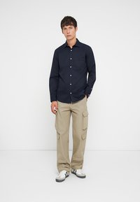 Jūras spēku krekls ar pogām un krūšu kabatu, kombinēts ar bēšiem cargo biksēm ar vairākiem kabatām, un baltiem sporta apaviem ar melnām svītrām.