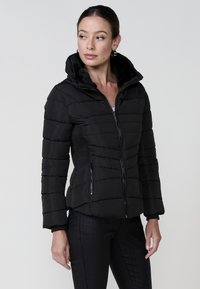 Chaqueta de plumas negra con un alto cuello, cierre frontal con cremallera y bolsillos laterales. Presenta un diseño acolchado y un forro interior suave.