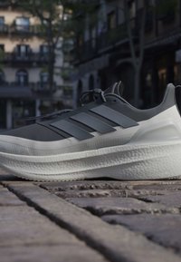 Σκούρο γκρι και άσπρο αθλητικό παπούτσι Adidas με παχιά σόλα που ακουμπά σε λιθόστρωτο δρόμο σε αστικό περιβάλλον με θολό φόντο κτιρίων.