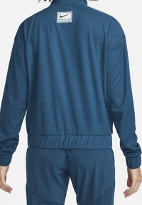 Blå Nike Team jakke med høj krave, elastiske manchetter og taljebånd. Har en lille logo-patch på øverste del af ryggen. Glat stoftekstur.