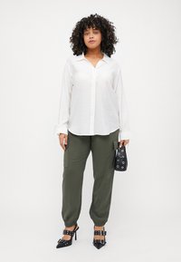 Λευκό πουκάμισο με μακριά μανίκια, ελαιόχρωμο παντελόνι cargo και μαύρες ψηλές γόβες με τσιμπιδάκια. Κρατά μια μικρή μαύρη τσάντα χειρός.