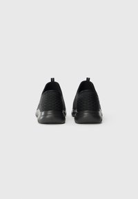 Pair of black slip-on sneakers with textured back panels and pull tabs, shown from the rear on a light gray background.