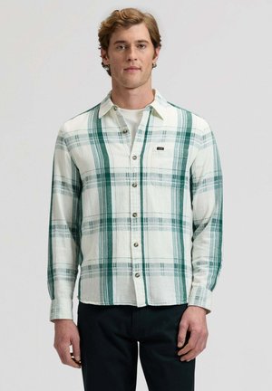 Młody mężczyzna w białej i zielonej kraciastej koszuli zapinanej na guziki, na białej koszulce, ubrany w czarne spodnie, stojący na tle szarego tła.