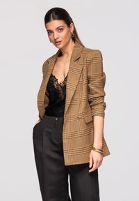 Kvinde iført brunternet blazer over sort blonder top og mørke bukser med høj talje, med guldringeøreringe og armbånd, stående foran en ensfarvet baggrund.