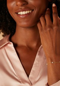 Goldene Armbanduhr mit zwei Perlenakzenten, getragen an einer Hand mit glatter Textur. Das Modell trägt eine hellrosa Satinbluse.