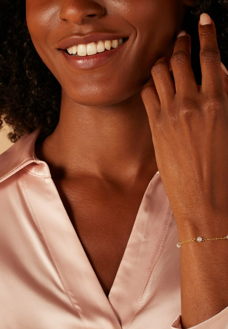 Goldene Armbanduhr mit zwei Perlenakzenten, getragen an einer Hand mit glatter Textur. Das Modell trägt eine hellrosa Satinbluse.