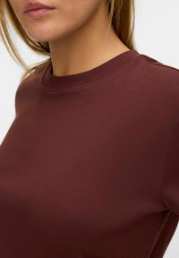 bordowa bawełniana koszulka z okrągłym dekoltem o gładkiej fakturze, z prążkowanym kołnierzem i dopasowanym krojem, pokazująca zbliżenie na linię szyi i ramię.