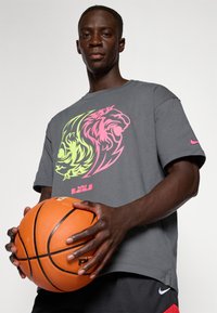 Grå bomulds t-shirt med et stort flerfarvet lionsgrafik i pink og grøn. Holder en orange basketball. Sorte shorts med røde accenter.