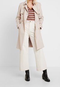 Kvinna iklädd beige trenchcoat, randig v-ringad topp, högmidjade vida vita byxor och svarta ankelboots, stående med händerna i fickorna.