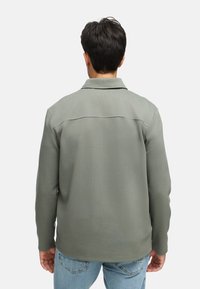 Olivengrønn langermet skjorte med teksturert stoff, krage og rett nederkant, kombinert med lyseblå jeans. Sett bakfra.