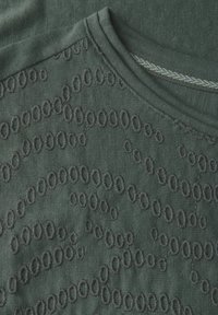 Un tissu vert foncé présente un design texturé avec des motifs circulaires brodés superposés sur une base lisse. Le col présente un détail de couture contrasté.