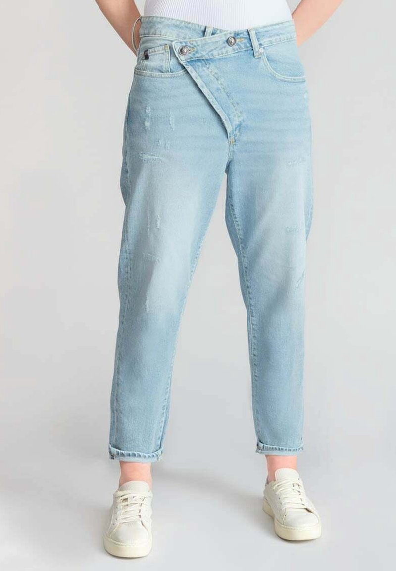 Vaqueros de mezclilla azul claro con un corte relajado, detalles desgastados y diseño de cinturilla asimétrica, combinados con zapatillas blancas.