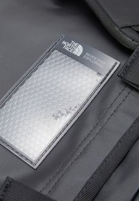 Svart fabric ryggsekk med et hexagon-mønstrete gjennomsiktig felt som viser merkevare og tekst. Metalliske detaljer og slitesterk søm er synlige.