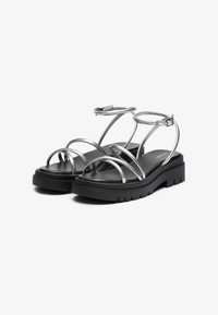 Sandalias metálicas plateadas con tiras y una suela gruesa negra, que presentan tiras delgadas, hebilla ajustable en el tobillo y una plantilla de textura suave.