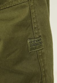Lähikuva oliivinvihreästä kankaasta, jossa on ommeltu etiketti, jossa lukee "G-STAR ORIGINALS RAW DENIM" pystysauman lähellä.