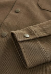 Tela de pana marrón con acanalado vertical, que presenta un puño plegable y botones de presión en tono plateado. Textura suave y detalles de diseño ajustados.