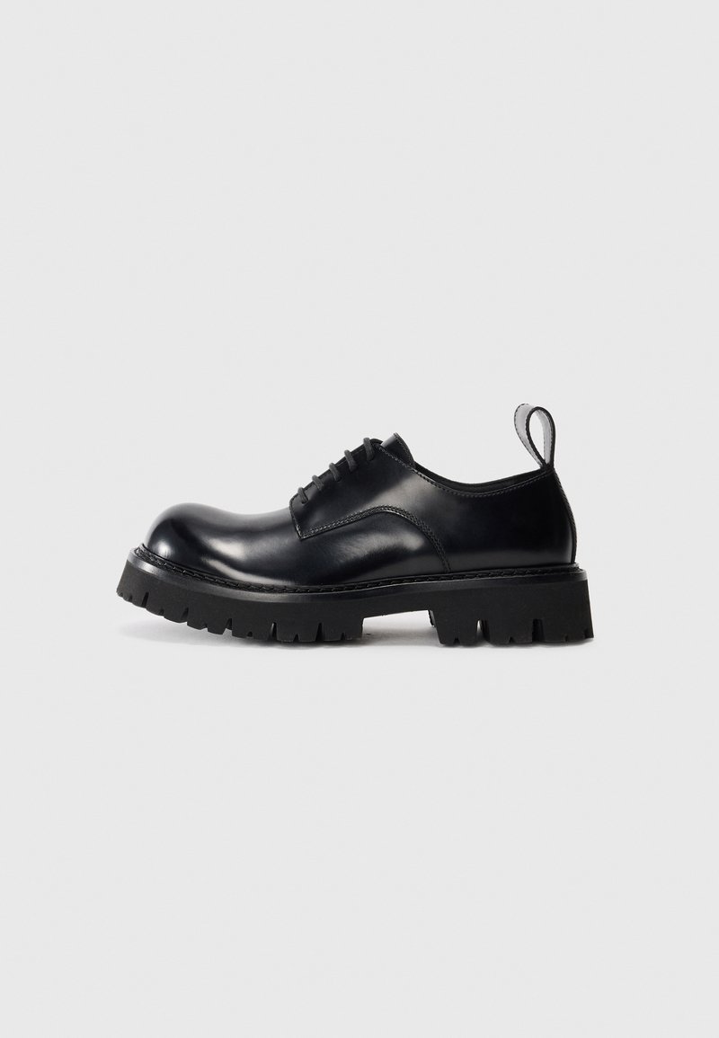 Black leather oxford shoe with a rounded toe, lace-up design, and a thick, textured rubber sole. Features a rear pull tab for ease.