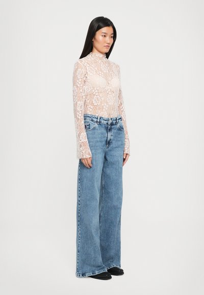 Top de encaje de manga larga en beige con patrón floral, combinado con jeans de pierna ancha de tiro alto en azul claro, que presentan un ajuste holgado y calzado oscuro.