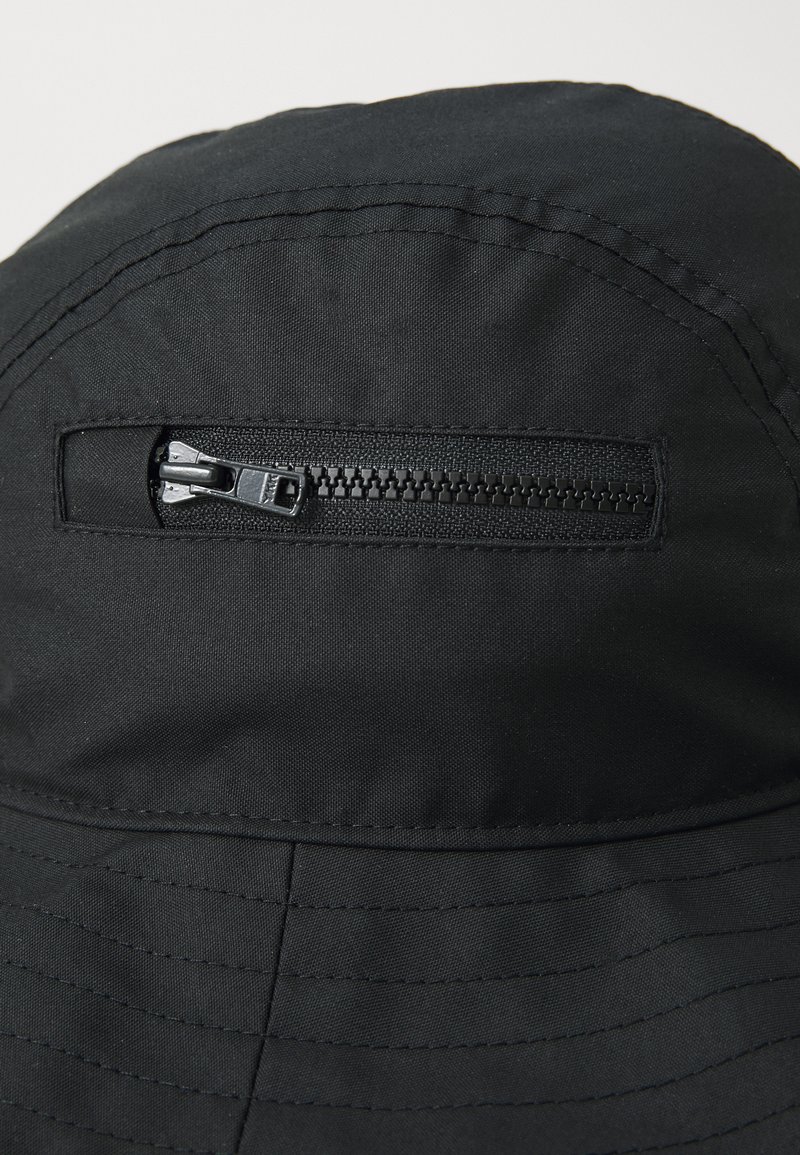 Sombrero tipo bucket negro hecho de un tejido texturizado con un detalle de cremallera horizontal en la parte delantera. Presenta líneas cosidas a lo largo de la base.