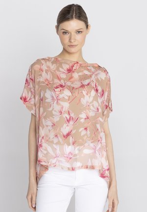 Kobieta ubrana w beżową przejrzystą bluzkę w kwiaty różowo-białe, do tego białe spodnie, stojąca na jednolitym tle.