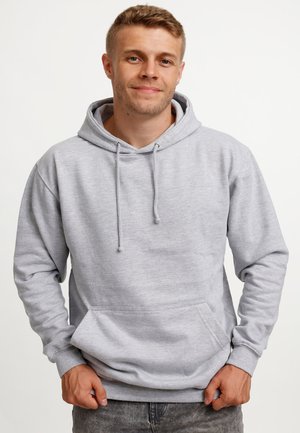 Młody mężczyzna z krótkimi włosami, ubrany w jasnoszarą bluzę z kapturem i ciemne dżinsy, stojący na jednolitym białym tle.