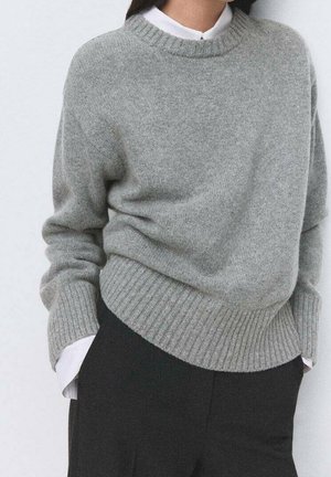 Personne portant un pull en maille gris sur une chemise blanche à col avec un pantalon noir, debout les mains dans les poches devant un fond uni.