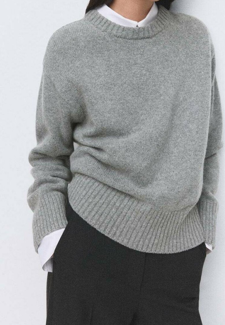 Personne portant un pull en maille gris sur une chemise blanche à col avec un pantalon noir, debout les mains dans les poches devant un fond uni.